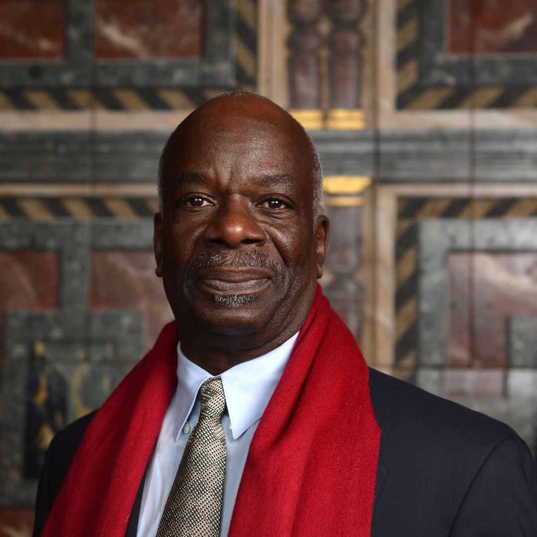 Joseph Marcel looks into the camera wearing a grey suit and red scarf