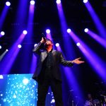 Shot from below of George Michael Impersonator in black suit and shirt singing, purple stage lights behind him