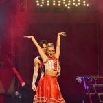 Blonde woman in crop top and long red skirt with blue fringing with her bare arms in the air, a brunet man leaning over her shoulder as they dance together.