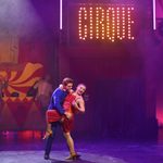 Blue suited white man holds dips red-dress wearing white woman who has her leg hooked on his waist as they dance.