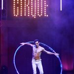 Bare-chested white brunet balances in a large hoop beneath lit up CIRQUE.