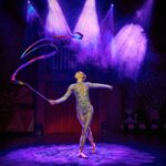 White woman in silver leotard twirls a rainbow ribbon to her right.