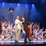 A group of people performing on stage with a boy and girl front and centre. Taken from Bugsy Malone, our Summer Youth Project 2013.