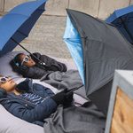 2 young girls laying outside on the floor wearing sunglasses and wrapped in coats sheltering from the rain with umbrellas.