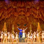 Genie (Yeukayi Ushe) and Aladdin (Gavin Adams) stand together, arms raised, on a golden platform in a golden room surrounded by golden dancers also with arms raised.