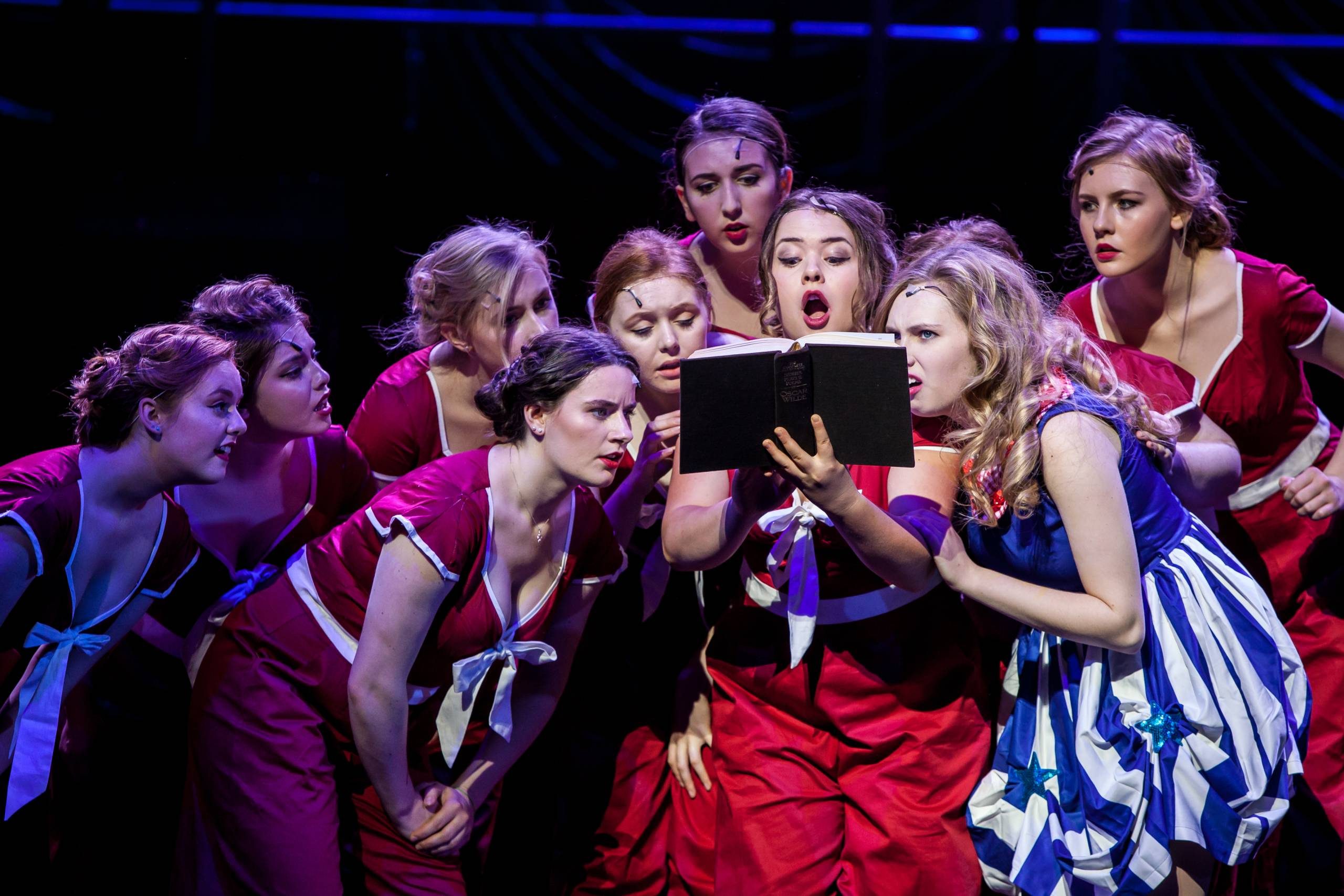 8 female members of the cast are standing around 1 cast member who is holding a black book wide open. They are gasping and look shocked.