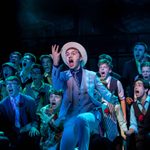 A young man in a 3-piece suit and hat sings and rolls dice in front of a group of young male actors from our Summer Youth Project 2017 - Guys and Dolls.