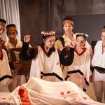 Northern ballet dancers in white tunics, flower crowns and round hats throwing rose petals over a bedsheet covered figure