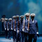 Seven soldiers carrying rifles stand behind each other with a blue and white lit stage.