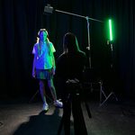 Young woman in white shirt and short check skirt standing in front of a green light taking direction from someone with their back to the camera in silhouette.