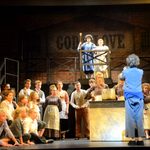 Young white male in blue trousers and blue jacket with his back to the audience holding a bowl begging for food whilst the other performers on stage stare at him.