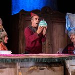 Three cast members sit at a table. Pinocchio in the middle holds a cake up to his face while the other two watch him.