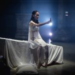 Juliet (Saeka Shirai) sitting on linen draped bench, wearing white and holding out a red vial