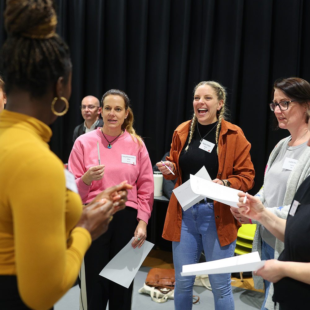 A diverse group of 5 female teachers stood in a circle talking and laughing amongst themselves.