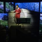 Danny Robins in long red raincoat standing on a platform outside a shed at night