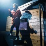 Danny Robins in long red raincoat walking down steps outside a shed at night