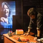 Danny Robins leans over a toy train circling on his wooden desk, staring intently at it. a white woman with curly grey hair projected on the wall