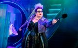 Ursula (Shawna Hamic) in purple denim jacket and glasses, holds a loo brush out, a urinal in the background.