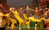 A group of young white dancers wearing yellow leggings and black and white checked coats with their right leg raised and their arms strecthed.