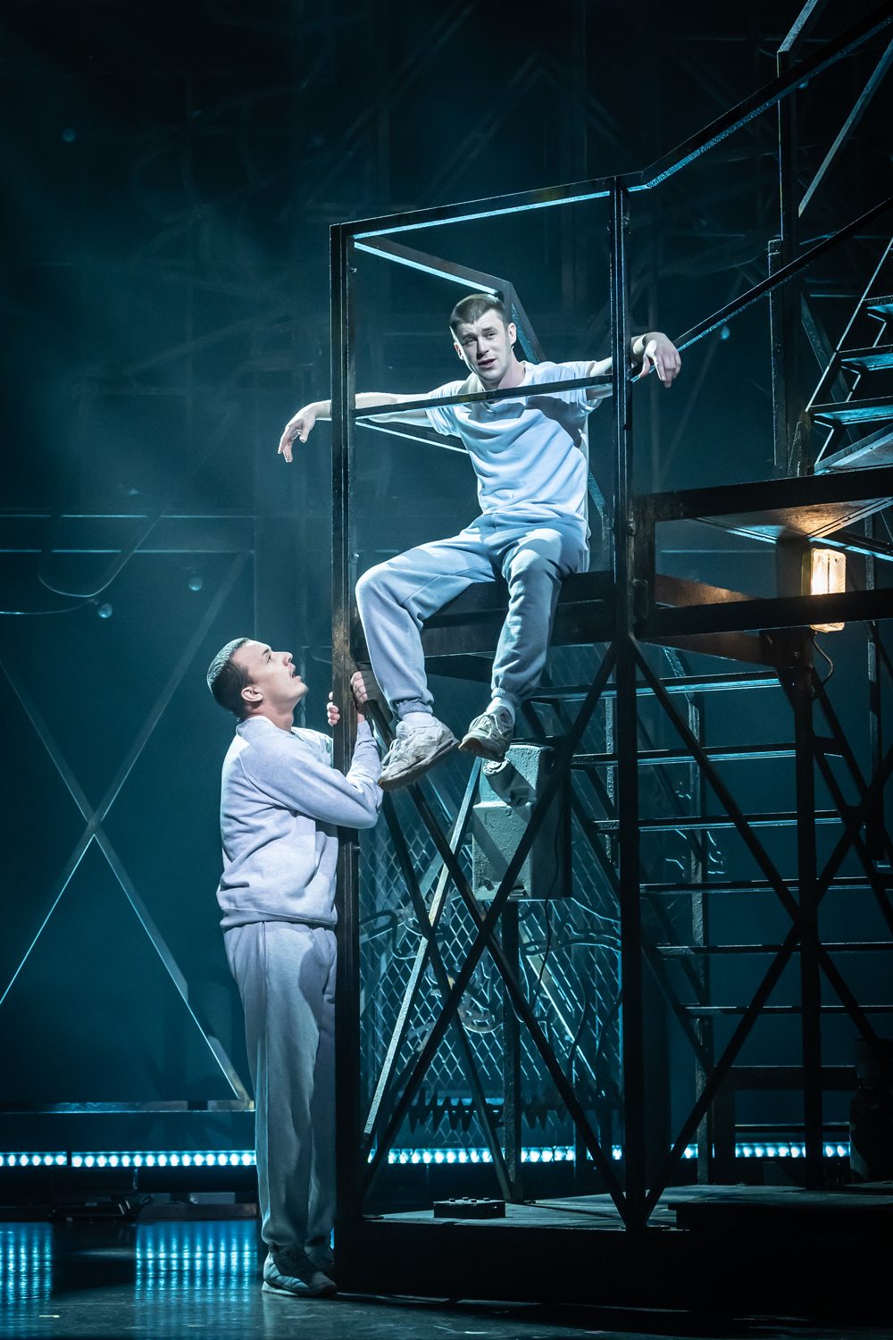 Sid Worley (Paul French) looks up at Zack Mayo (Luke Baker) sitting on metal stairs, both wearing grey tracksuits